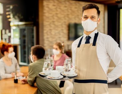 Happy waiter serving coffee to his customers while wearing protective face mask in a cafe due to coronavirus epidemic.