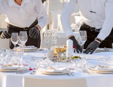 Close up waiters hands in protective black gloves arrange a wedding party reception table decorated with flowers: plates, forks, knives and wine glasses.